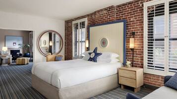 Habitación de hotel con una cama grande, un espejo redondo, un escritorio con lámpara y vistas al exterior mediante una ventana con persianas.