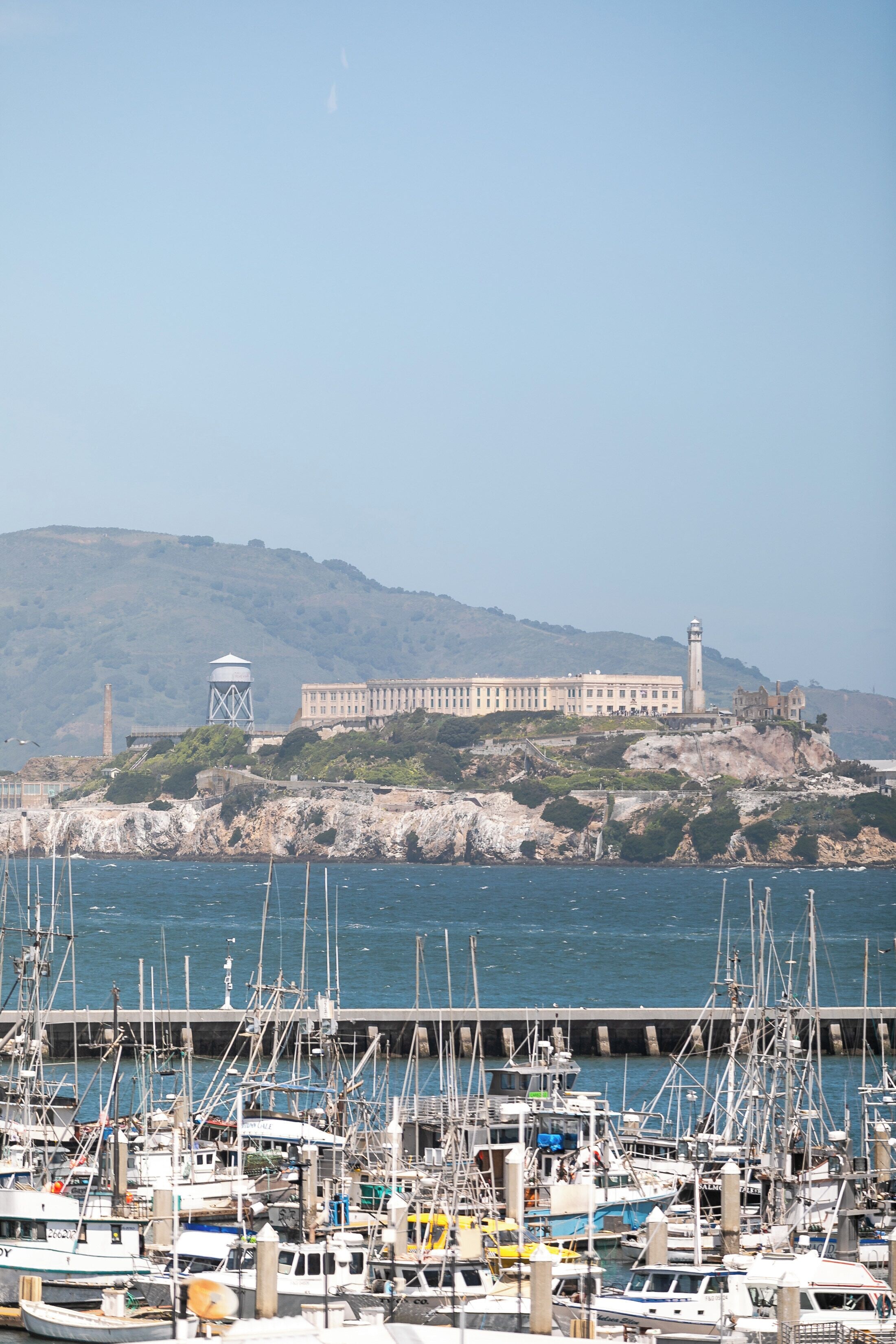 A marina with numerous boats docked, a lighthouse, and a large building on a hill in the background.