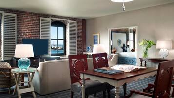 A hotel room with a brick wall, a large window with shutters, a dining table with chairs, a television, and a bed with a blue and white striped bedspread.