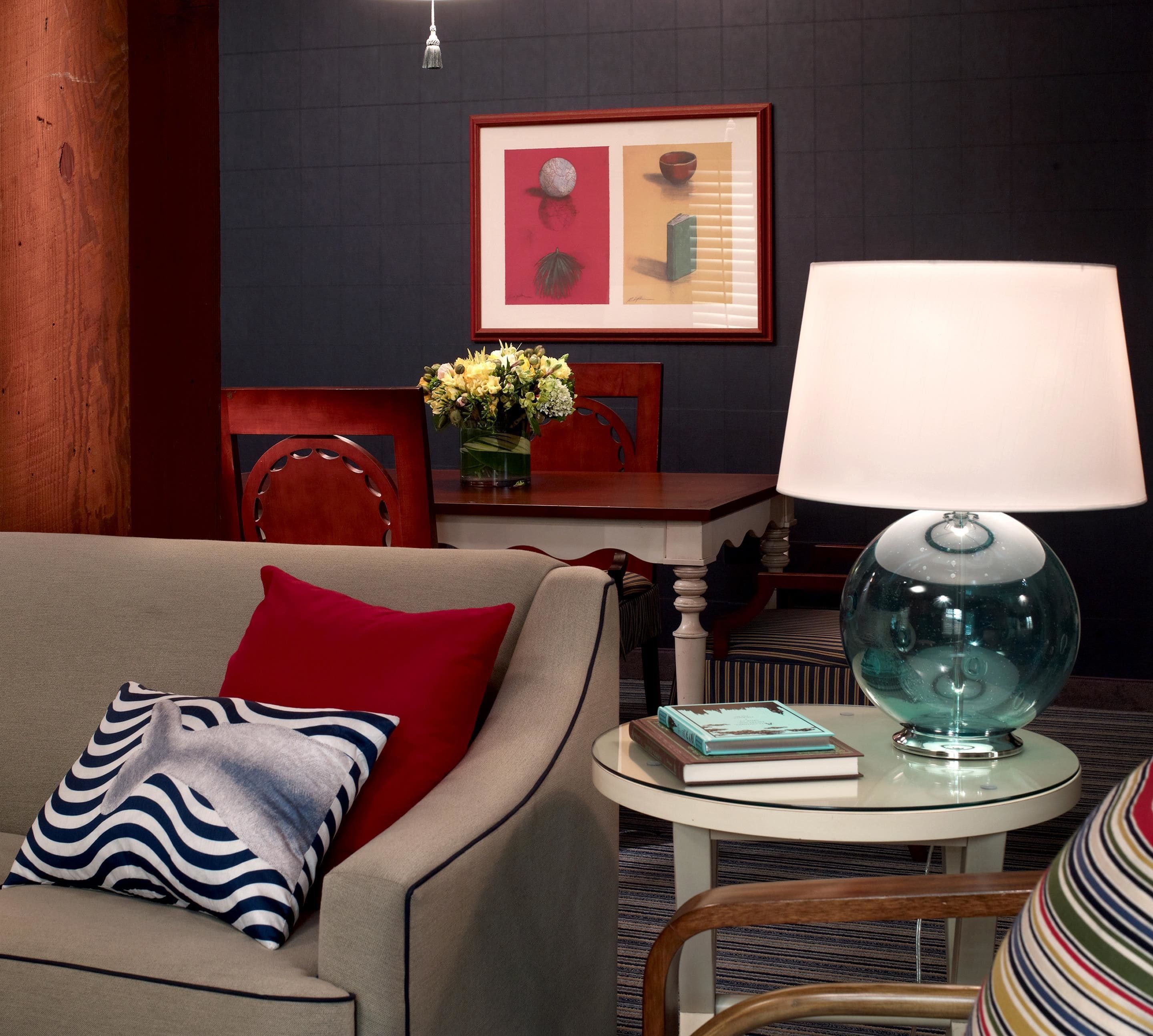 A cozy living room with a sofa, a round table with a lamp, a chair, and a framed artwork on the wall.