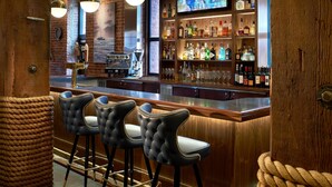 Un espace bar comprenant trois tabourets noirs, un comptoir en bois et une étagère remplie de bouteilles d’alcool.