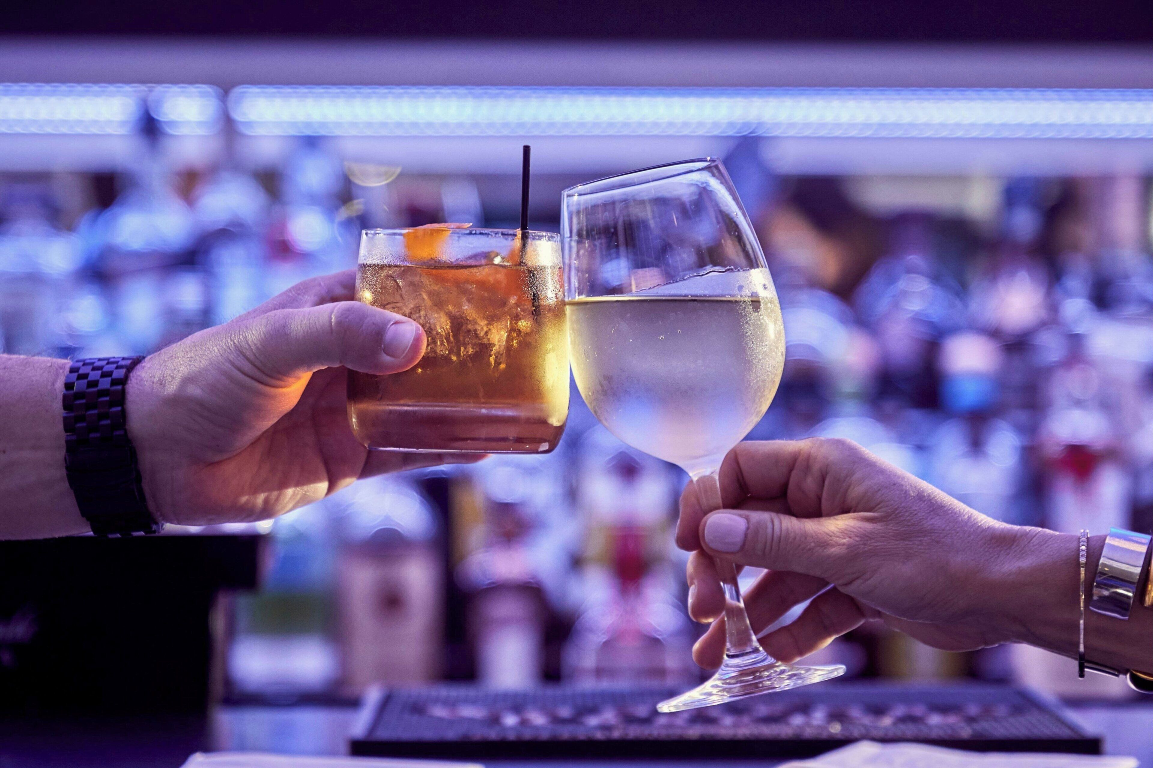 Duas mãos segurando copos com bebidas diferentes, possivelmente em um bar, com um fundo desfocado de garrafas.