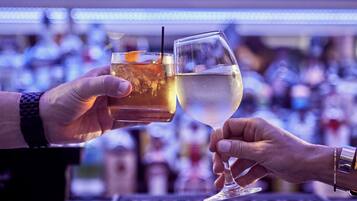 Dos manos sosteniendo vasos con bebidas diferentes, probablemente en un bar, con un fondo borroso de botellas.