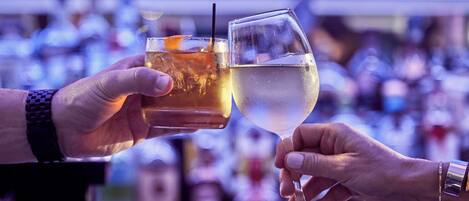 Two hands holding glasses of different beverages, likely in a bar setting with a blurred background of bottles.