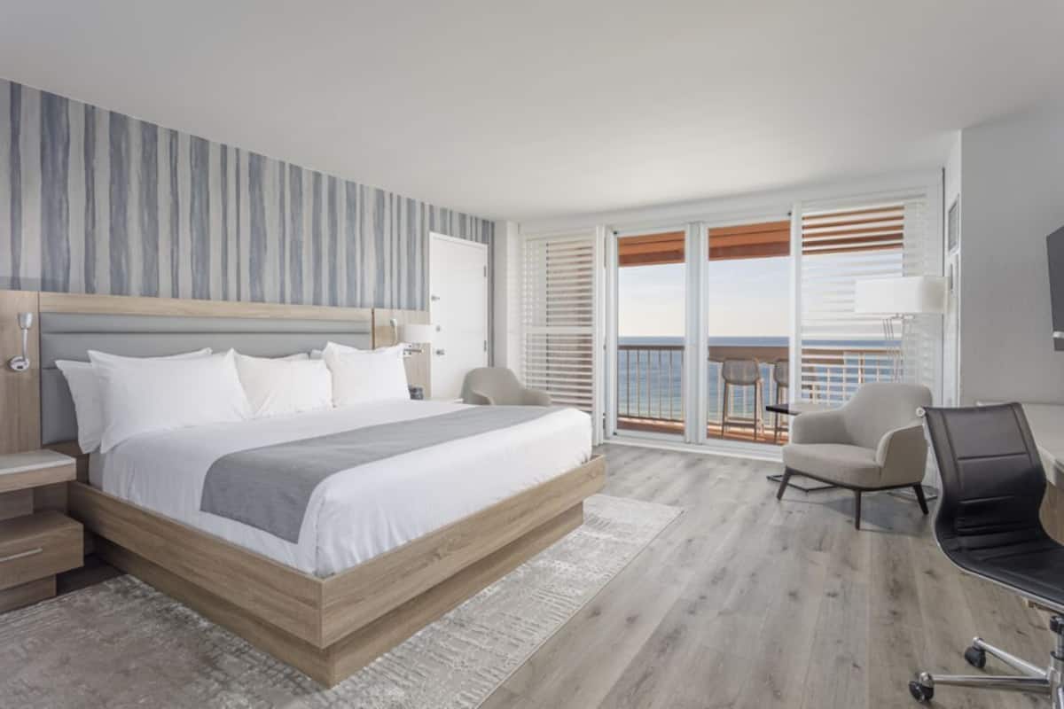 A hotel room with a bed, a chair, a desk, a balcony with a view, and a large window.