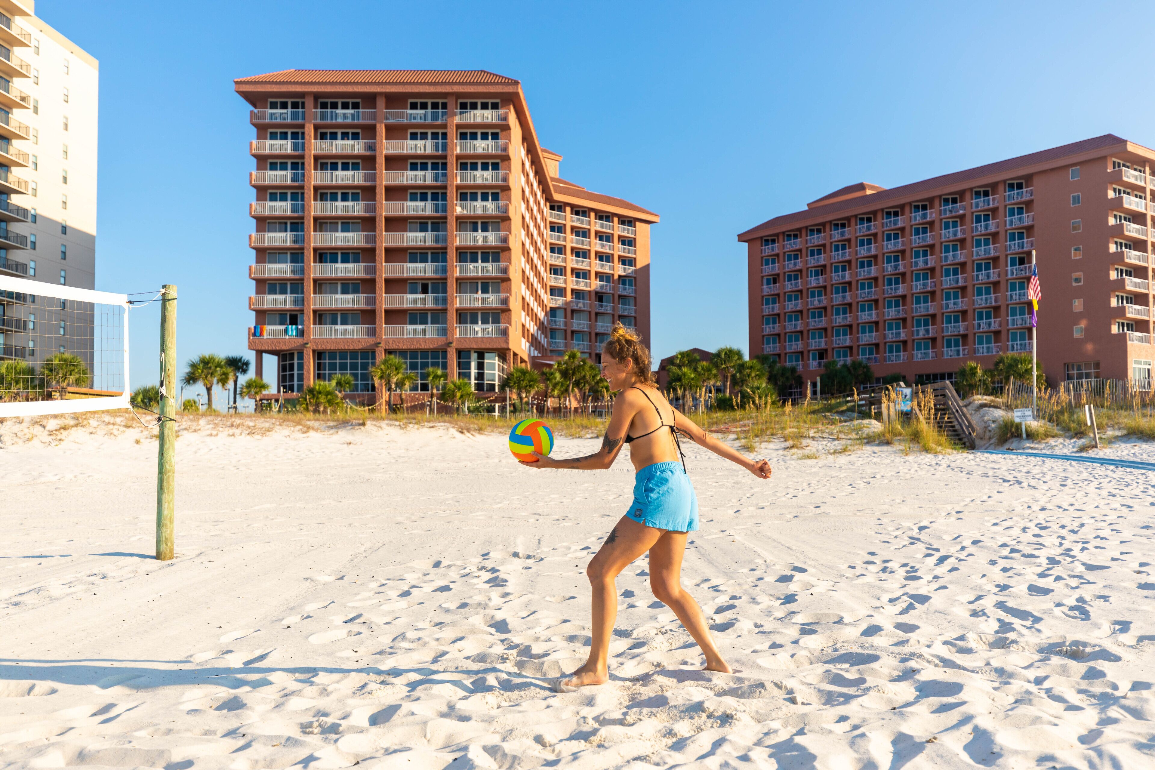Una playa con una persona jugando voleibol, arena y edificios altos al fondo.