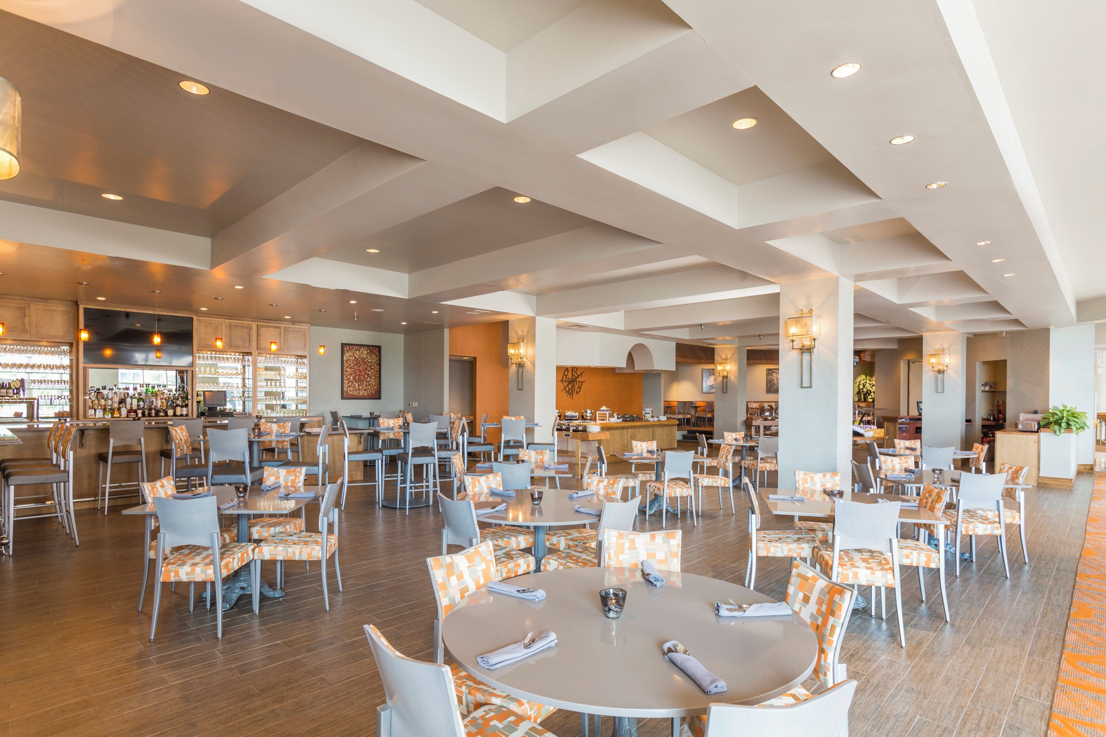 A spacious hotel lobby with multiple dining tables and chairs, a bar area, and a kitchen in the background.