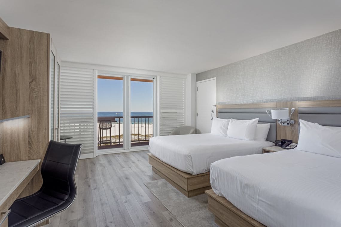 A hotel room with two beds, a desk, a chair, and a balcony with a view of the beach.