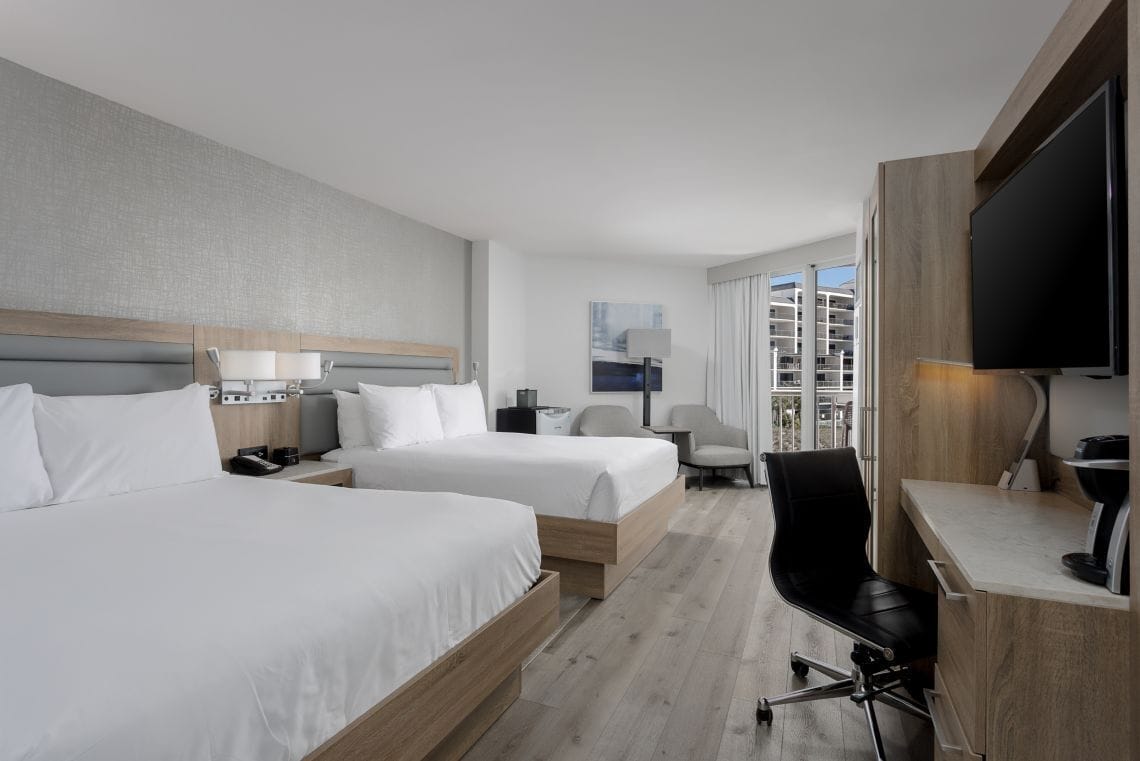 A hotel room with two beds, a desk, a chair, a TV, and a window with a view of buildings.