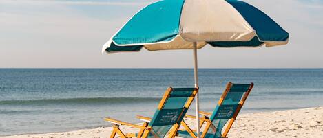 Une scène de plage avec deux chaises de plage en bois placées sous un parasol bleu et blanc, le tout sur une plage de sable avec l’océan en arrière-plan.