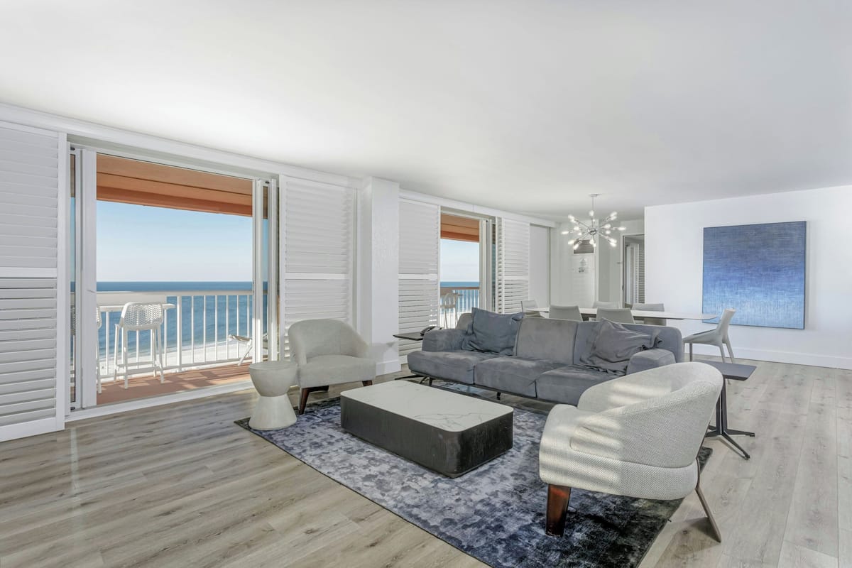 A modern living room with a grey sofa, white armchairs, a coffee table, and a view of the sea through large windows.