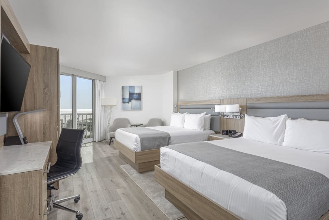 A hotel room with two beds, a desk, a chair, a TV, and a balcony with a view.