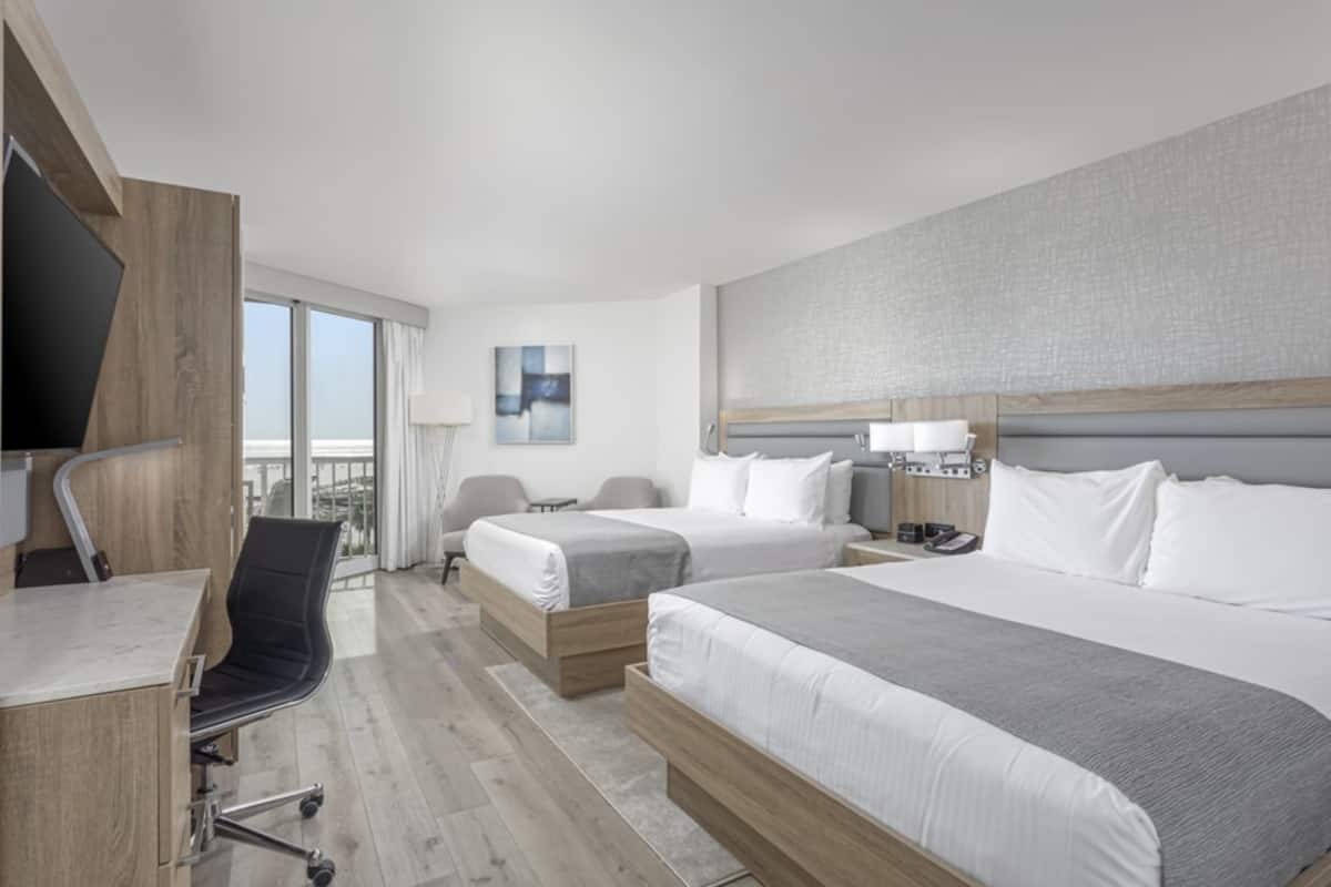 A hotel room with two beds, a desk, a chair, a TV, and a balcony with a view.