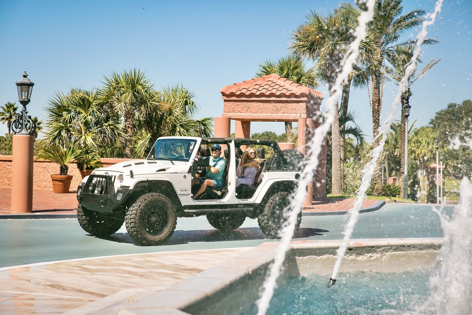 A white off-road vehicle parked near a fountain with a tiled walkway and palm trees in the background.