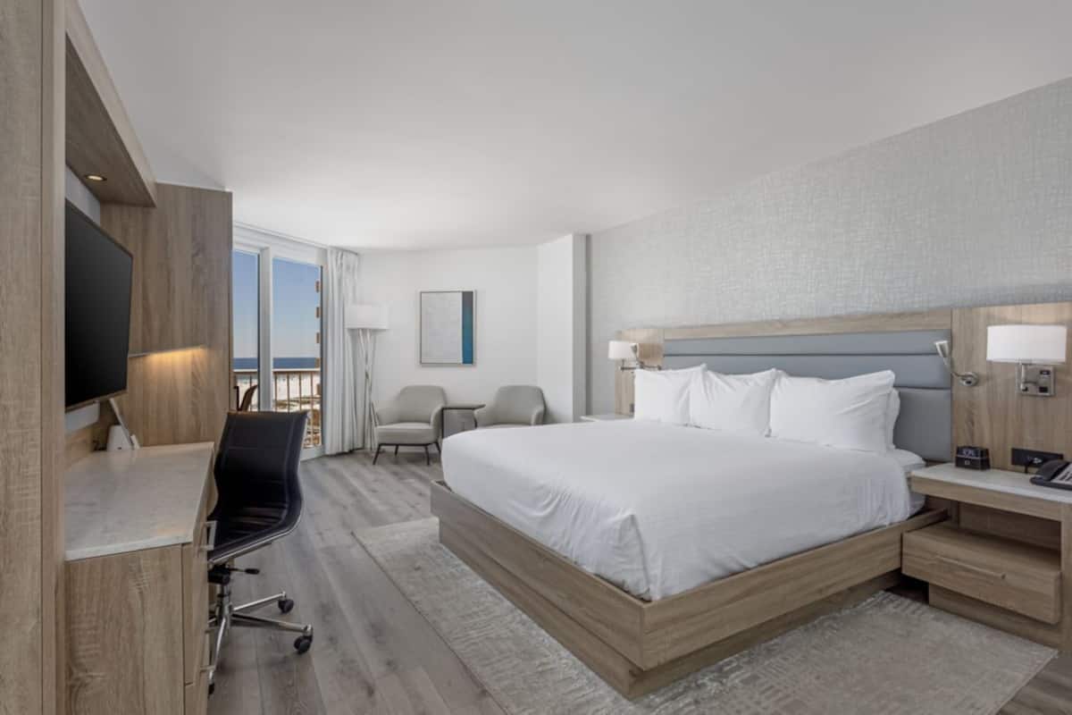 A modern hotel room with a large bed, a desk with a flat-screen TV, a chair, and a nightstand with a lamp.