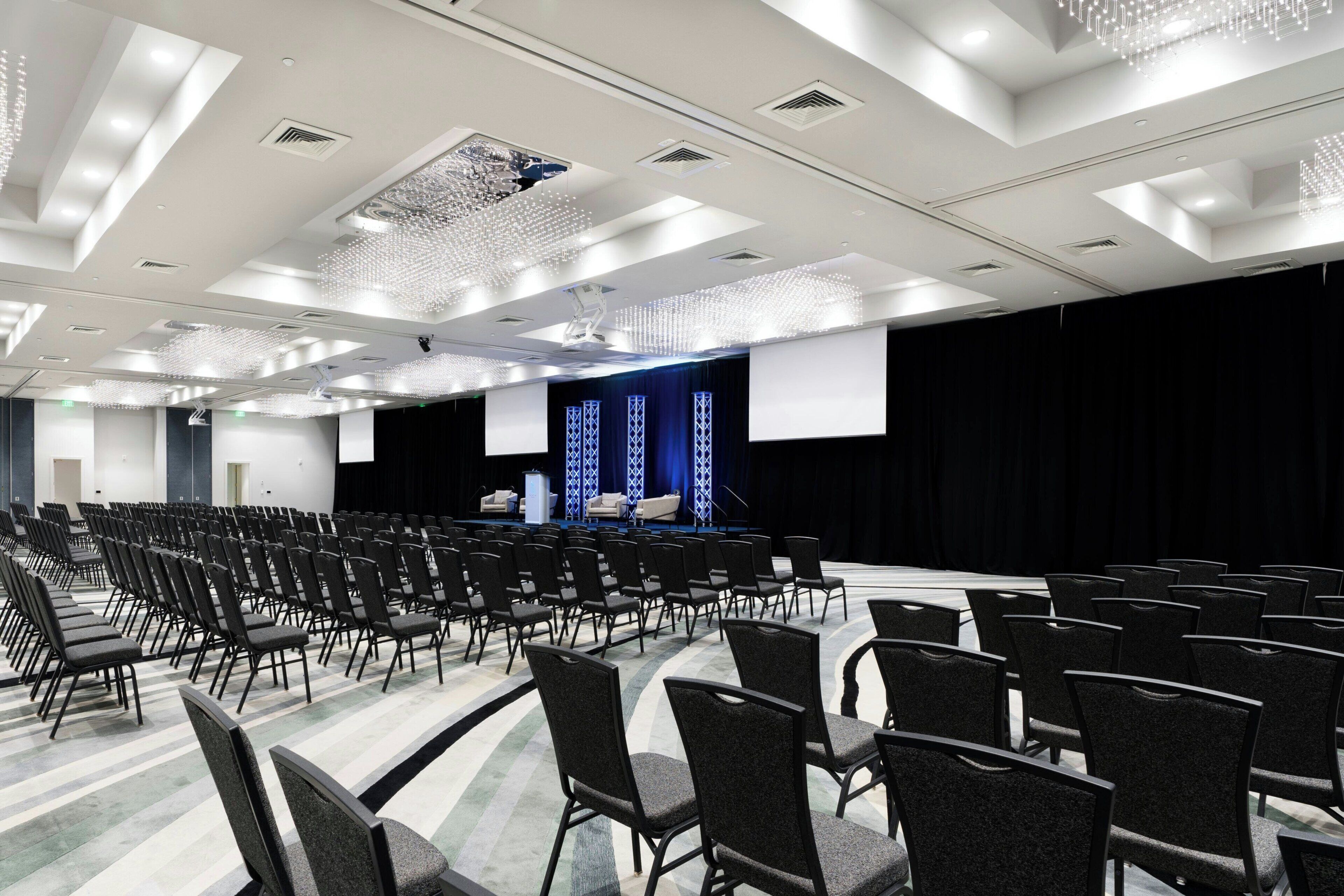 A conference room with rows of chairs, a stage with a podium, and a large screen.