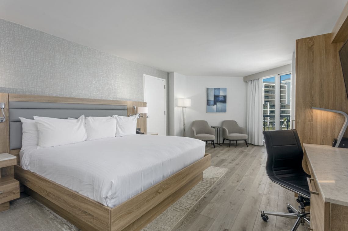 A hotel room with a bed, bedside tables, a desk, a chair, a lamp, and a view of the outside.