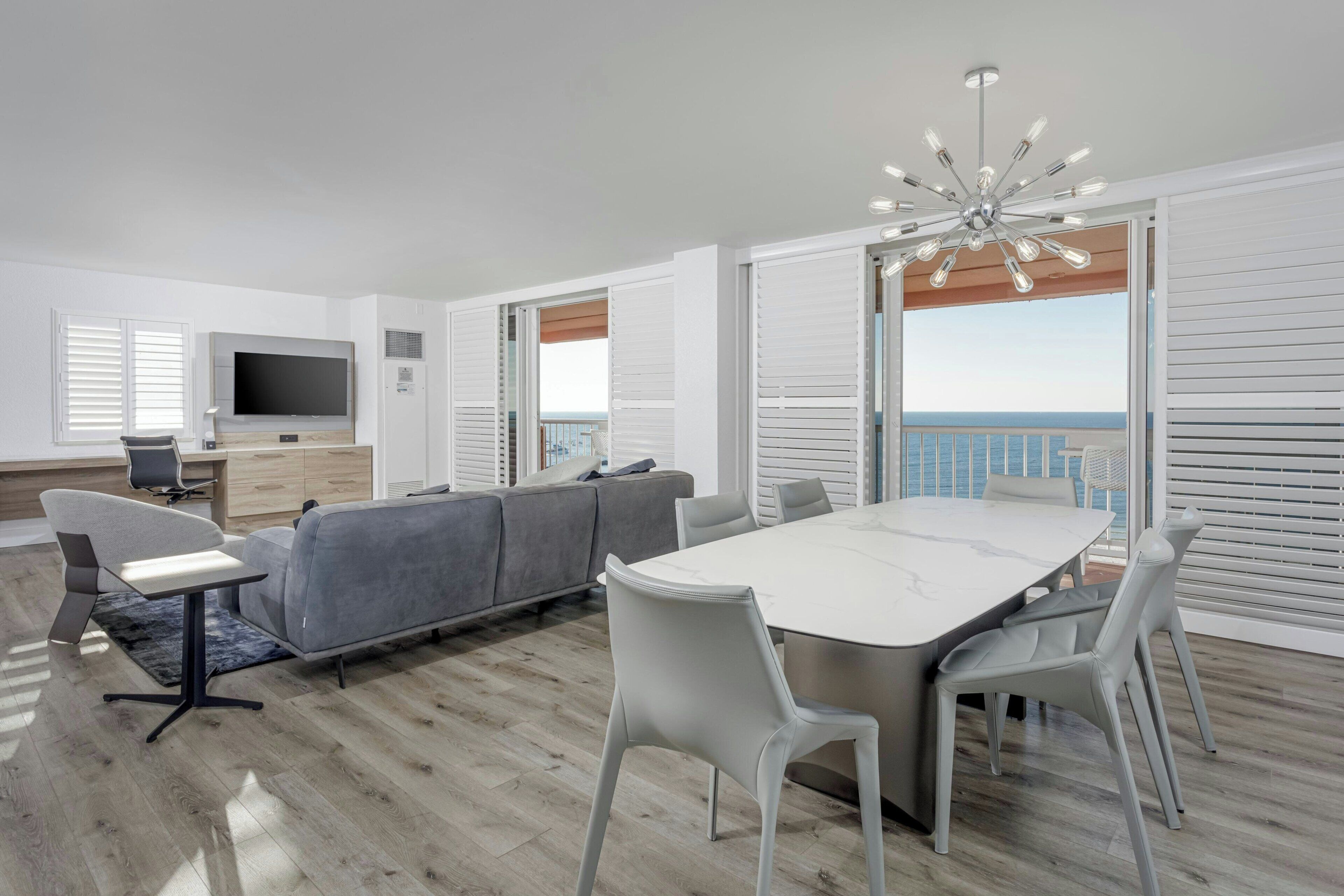 A modern living room with a grey sofa, a dining table with chairs, a TV, and a balcony with a view.