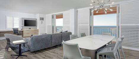 A modern living room with a grey sofa, a dining table with chairs, a TV, and a balcony with a view.