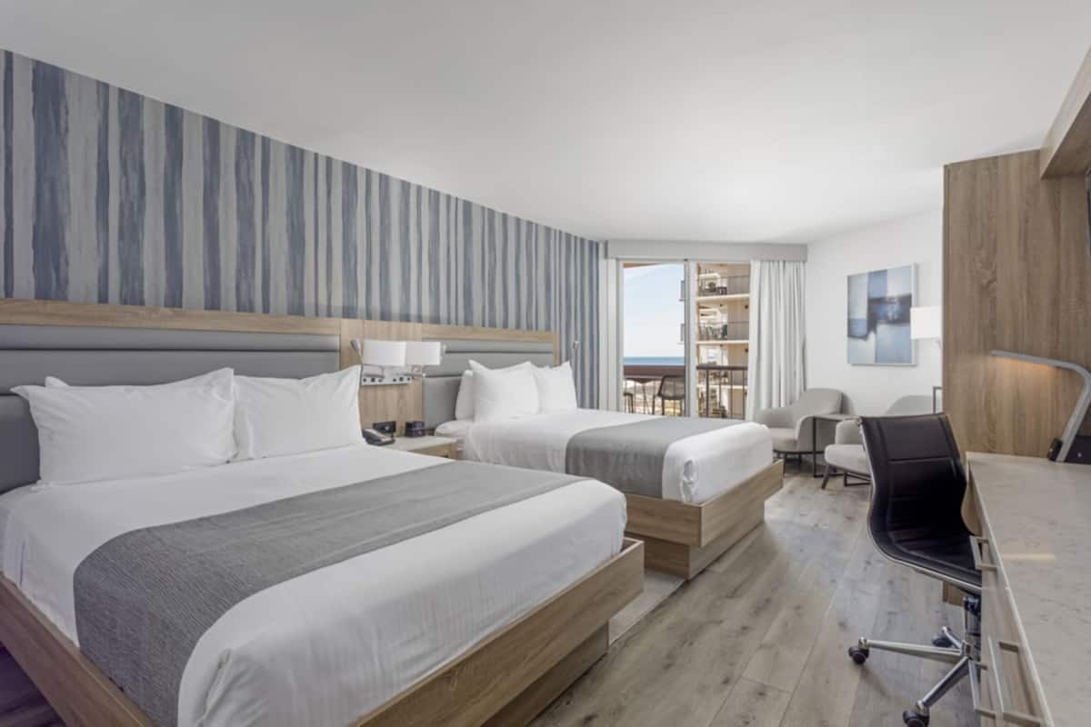 A hotel room with two beds, a desk, a chair, a bookshelf, and a view of the sea.