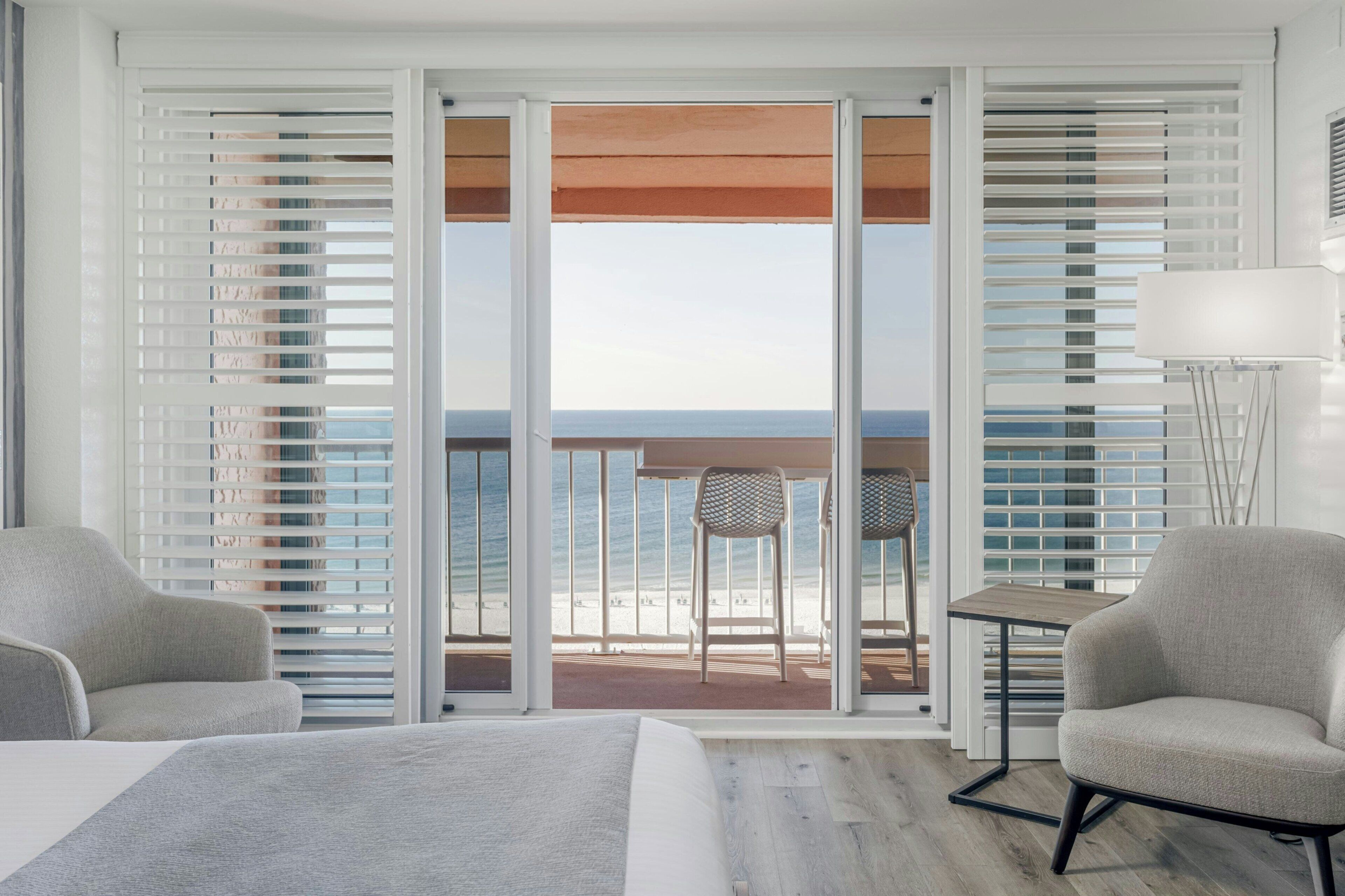 A hotel room with a bed, two chairs, a small table, and a view of the beach through a sliding door.