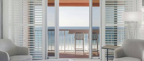 A hotel room with a bed, two chairs, a small table, and a view of the beach through a sliding door.