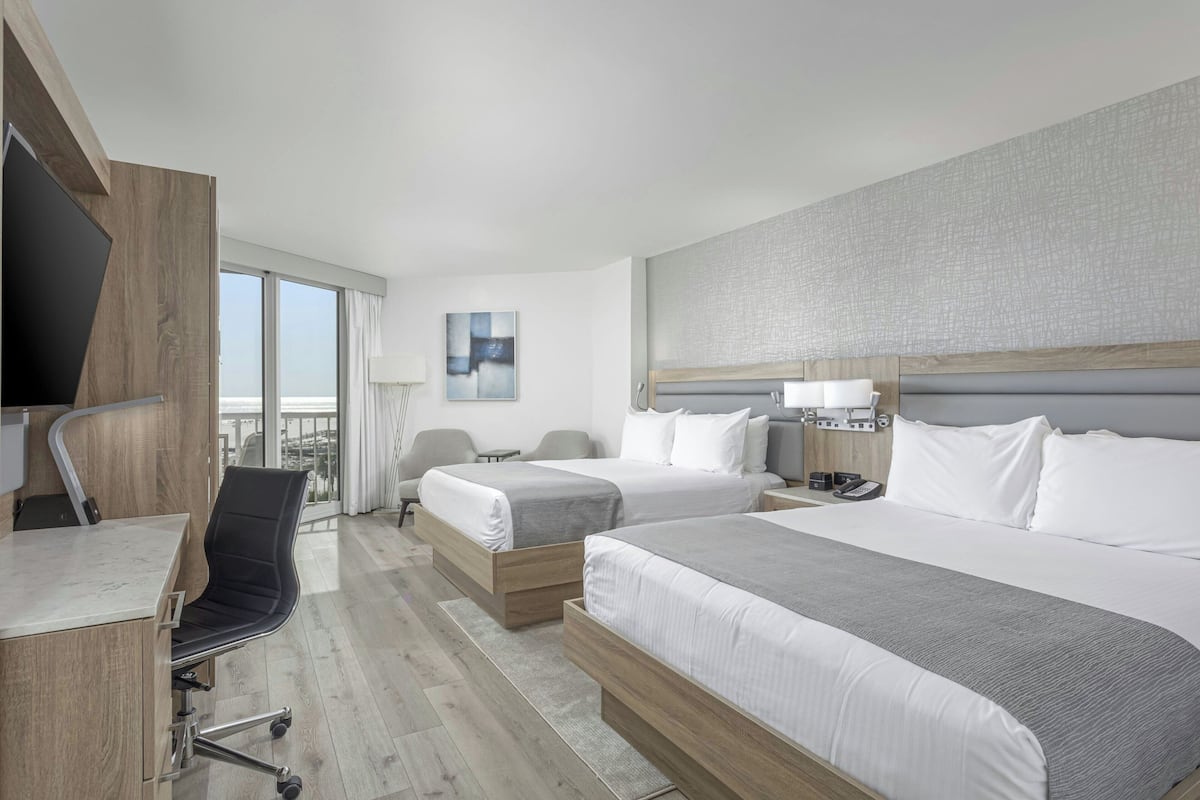 A hotel room with two beds, a desk, a chair, a TV, and a view of the city.