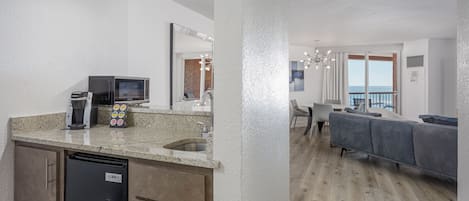 A modern kitchen with a built-in microwave, coffee maker, and refrigerator. A dining area with a gray sofa and a view of the ocean is visible through large windows.