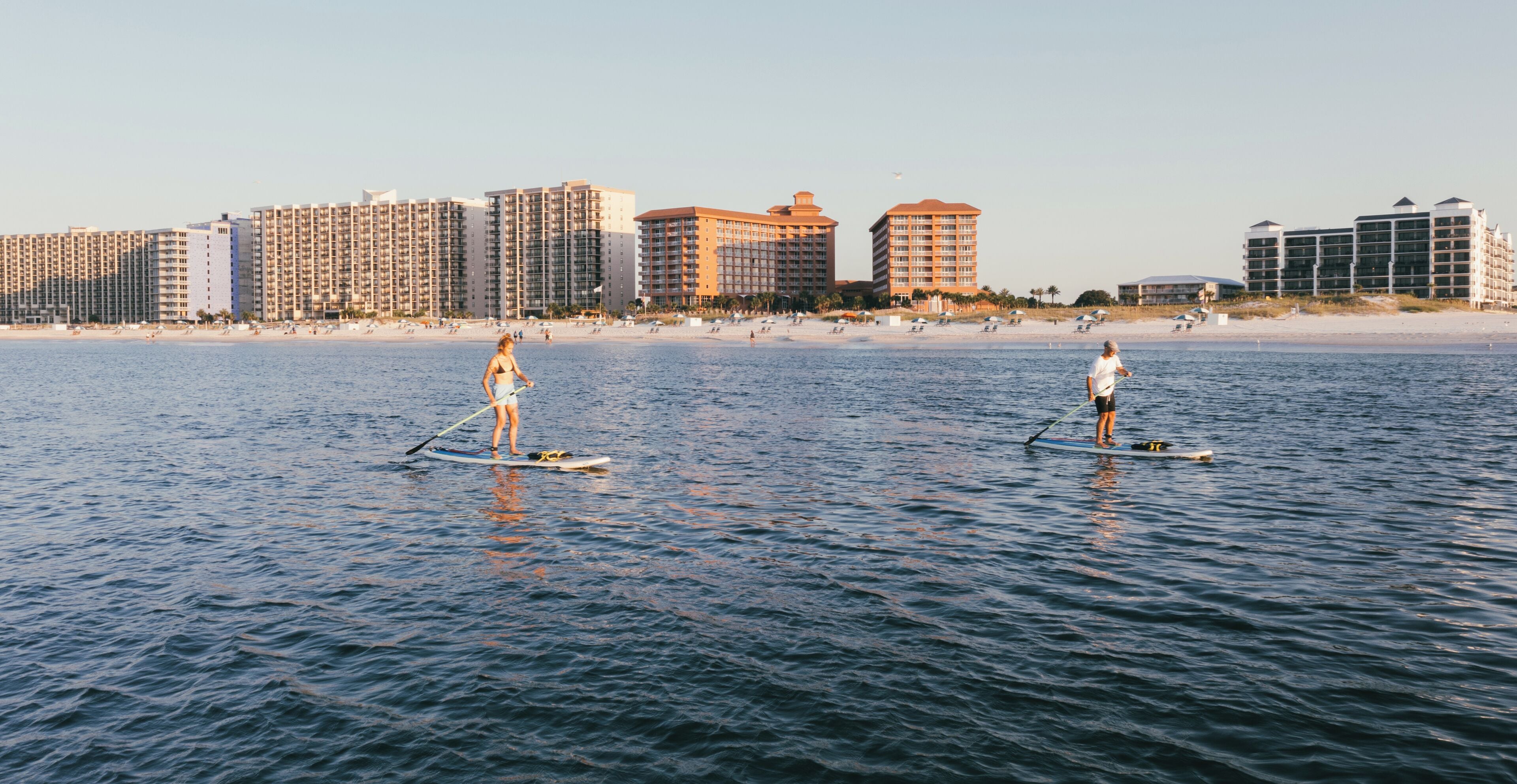En strand med paddleboardere og en række høje hotelbygninger i baggrunden.