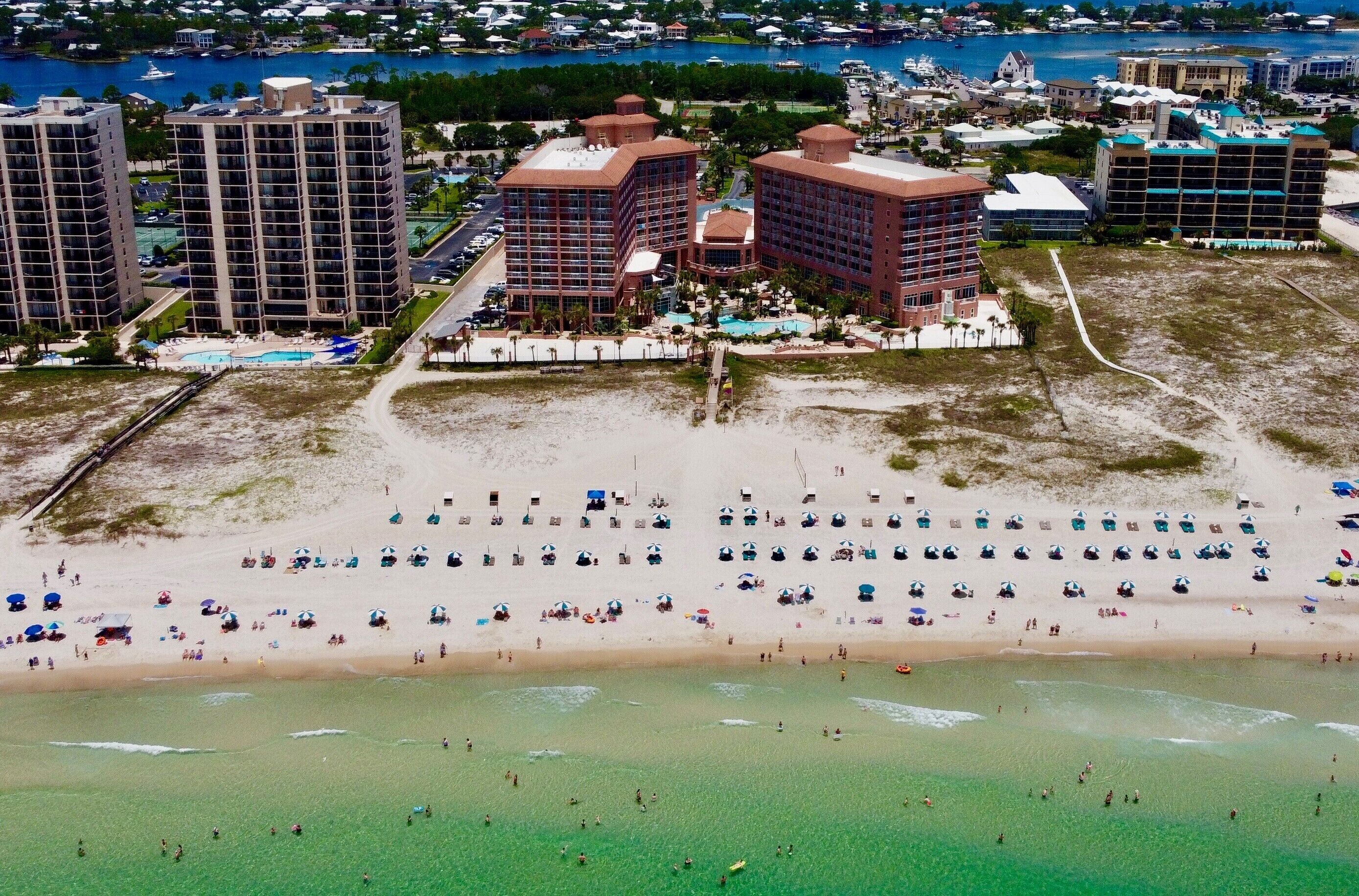 A coastal hotel with multiple high-rise buildings, a sandy beach with numerous umbrellas, and a clear view of the ocean.