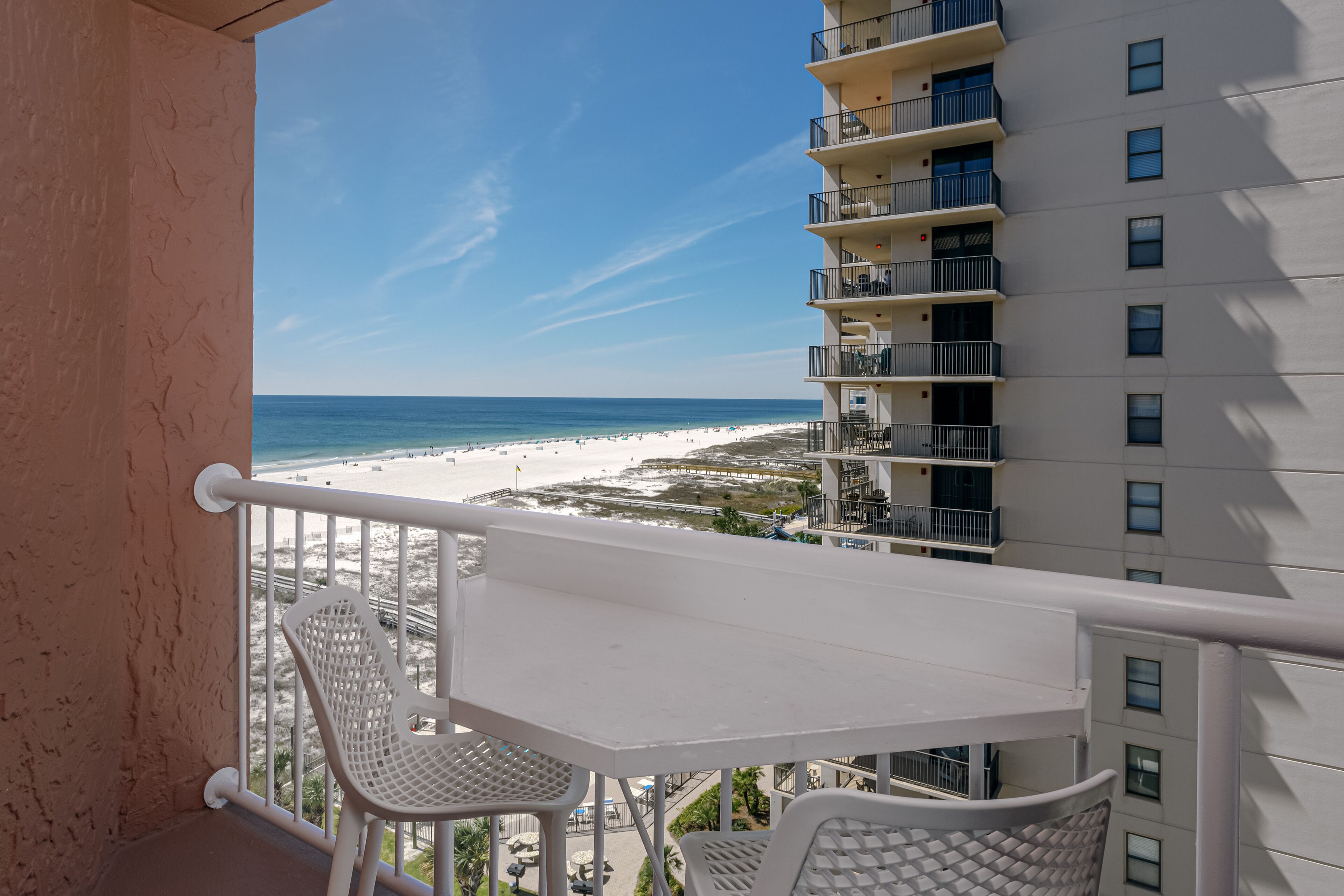 Un balcón con una mesa blanca y dos sillas, con vista a la playa y un edificio alto.