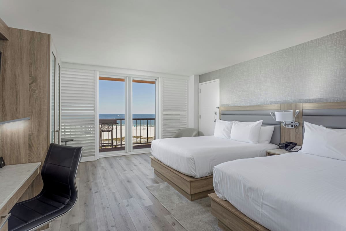 A hotel room with two beds, a desk, a chair, and a balcony with a view of the beach.