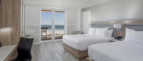 Et hotelværelse med to senge, et skrivebord, en stol og en balkon med udsigt til stranden.