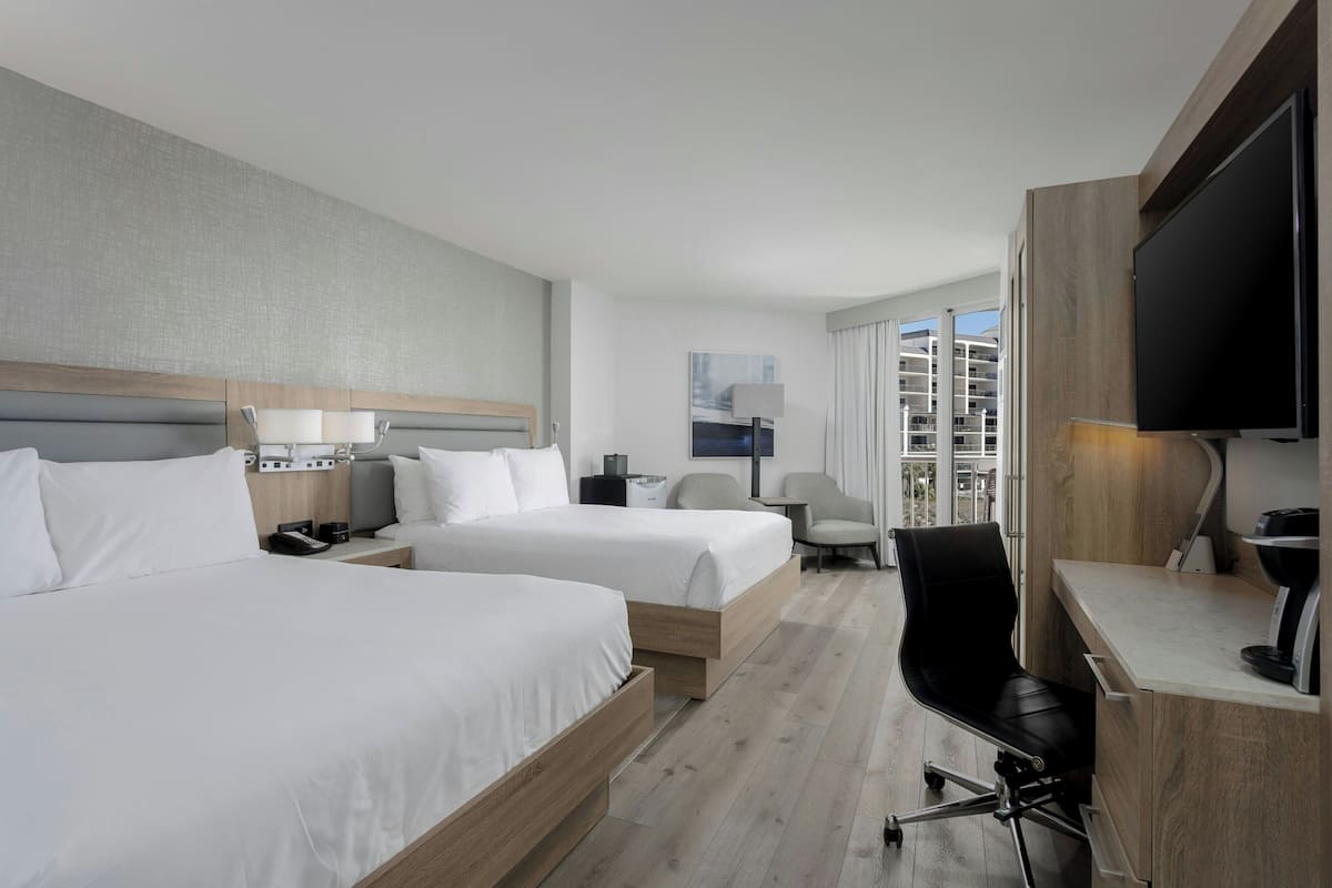 A hotel room with two beds, a desk, a chair, a TV, and a window with a view of buildings.