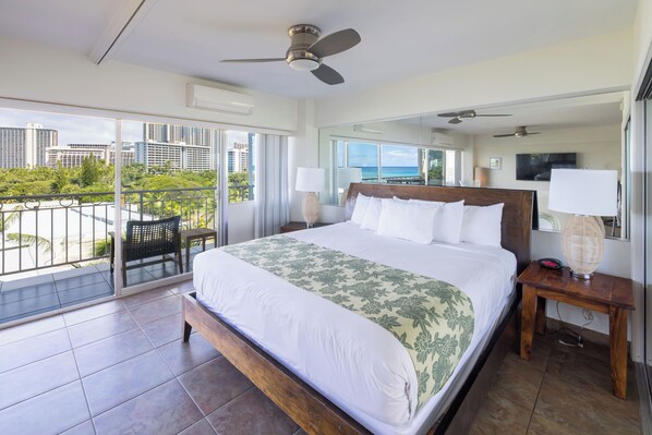 A spacious bedroom with a large bed, a balcony with a view of the city, and a ceiling fan.