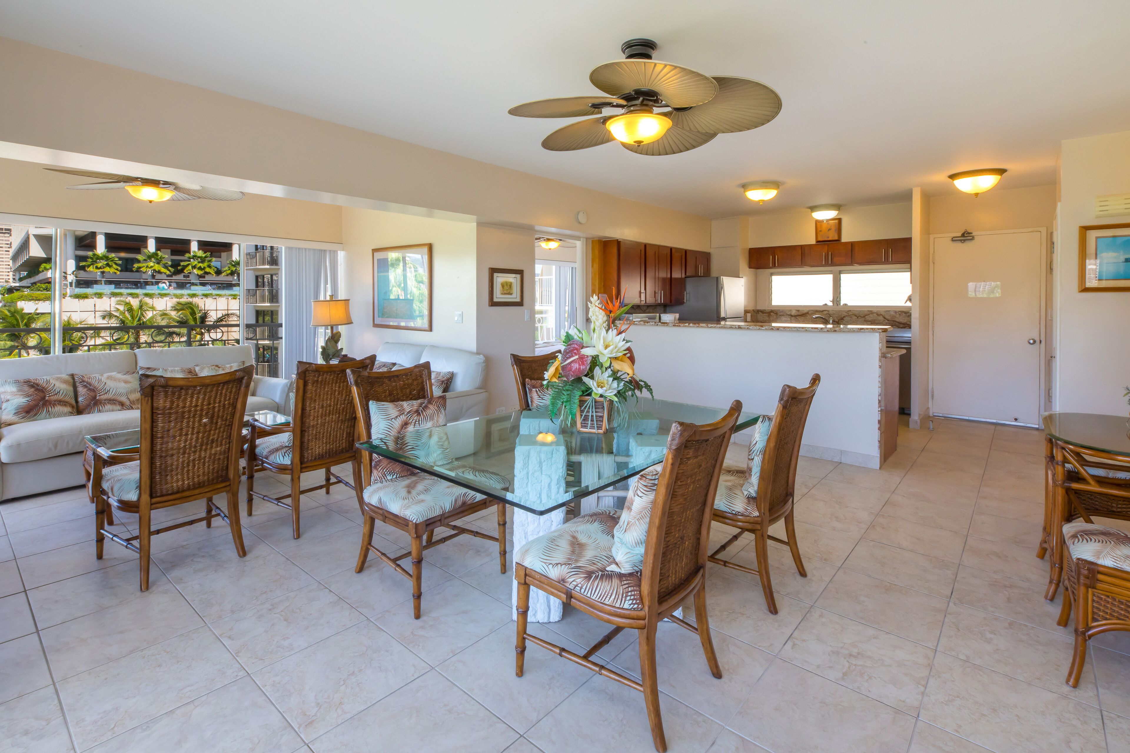 A spacious living area with a glass-top dining table surrounded by wicker chairs, a ceiling fan, and a view of a balcony with plants.