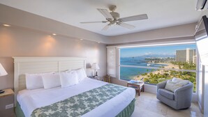 A hotel room with a large bed, a ceiling fan, a view of the ocean, a balcony, and a seating area.