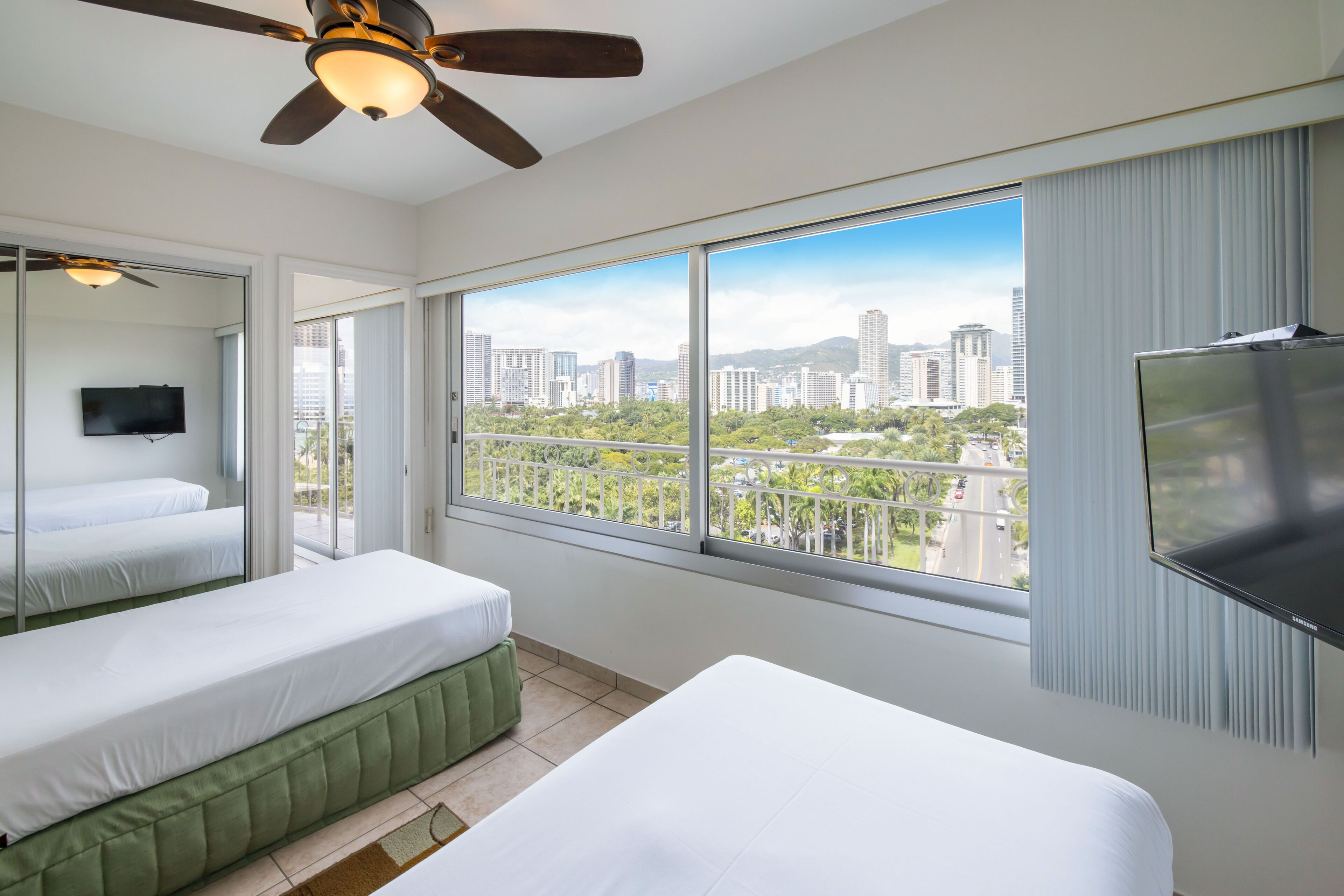 Ein Hotelzimmer mit zwei Betten, einem Deckenventilator, einem Fernseher und einem großen Fenster mit Blick auf die Stadt.