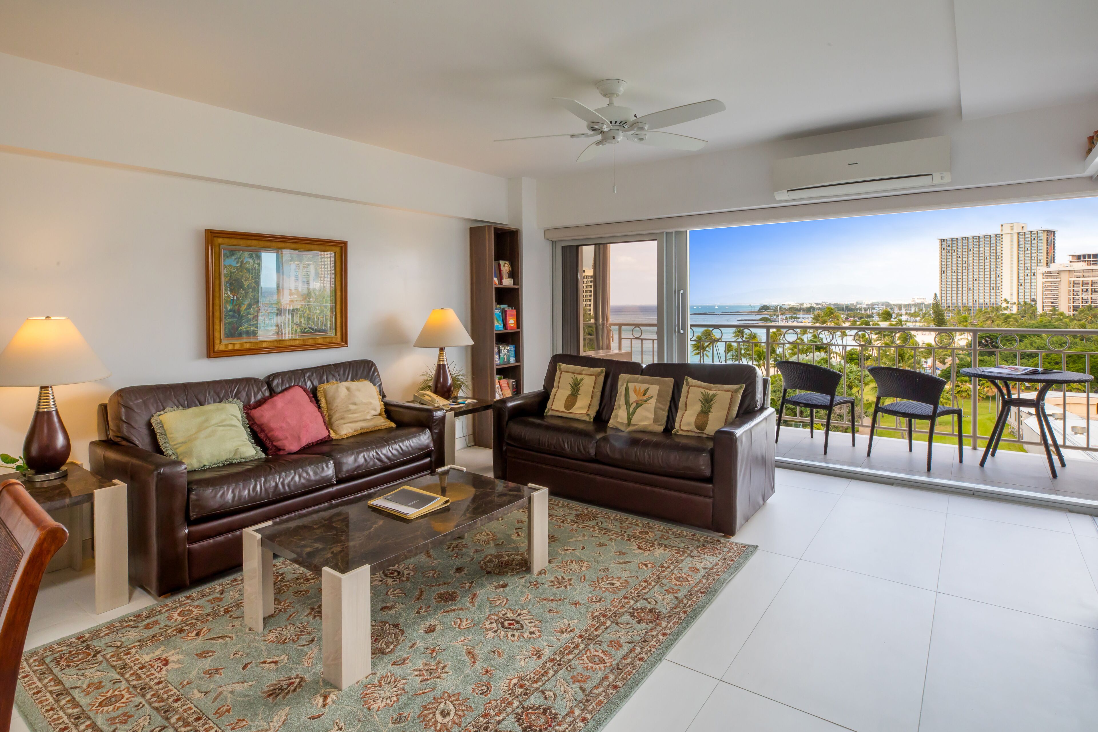 A living room with a sofa, coffee table, and chairs, a bookshelf, and a balcony with a view of the city and ocean.