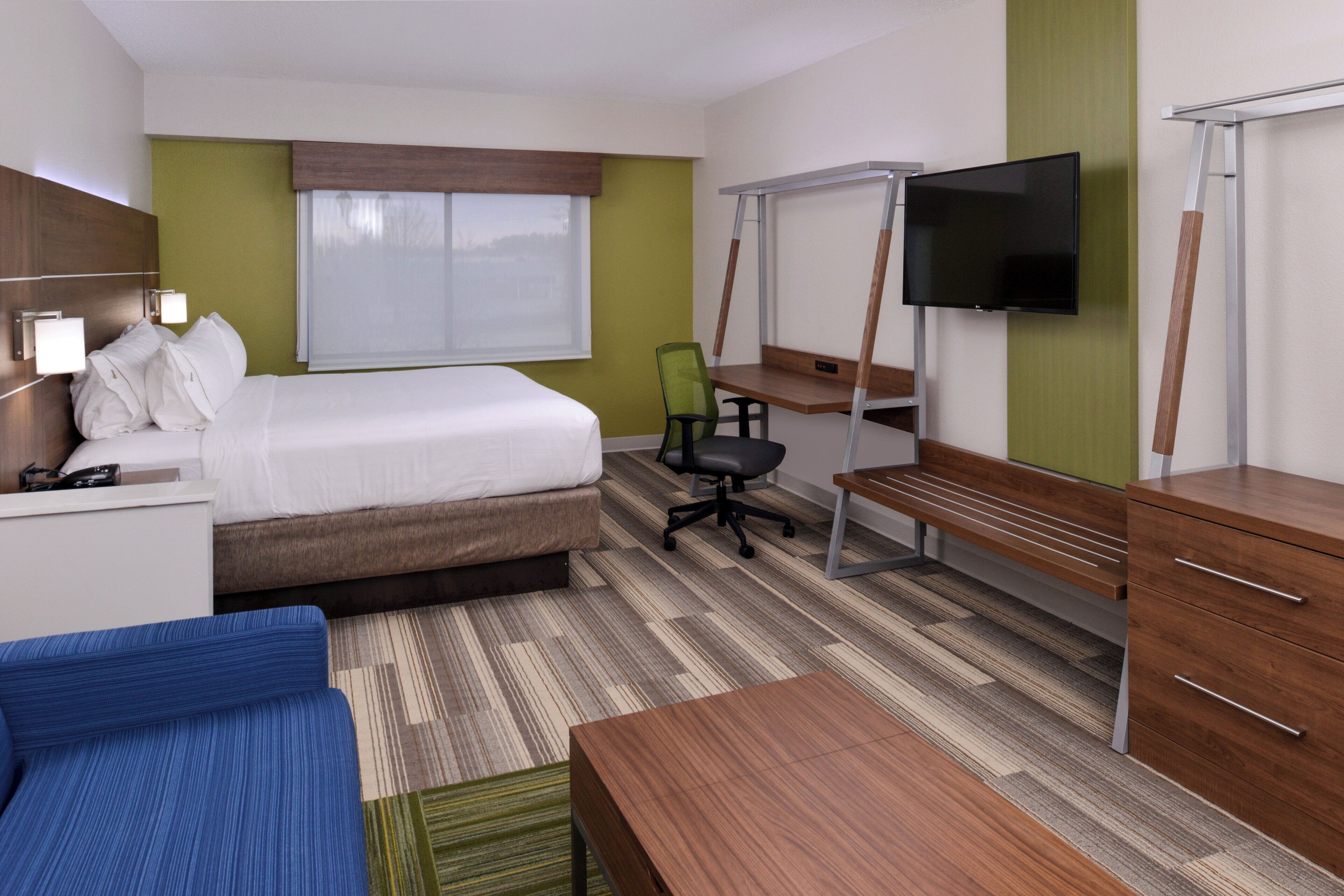 Quarto de hotel com cama, escrivaninha, cadeira, televisão e um pequeno sofá.