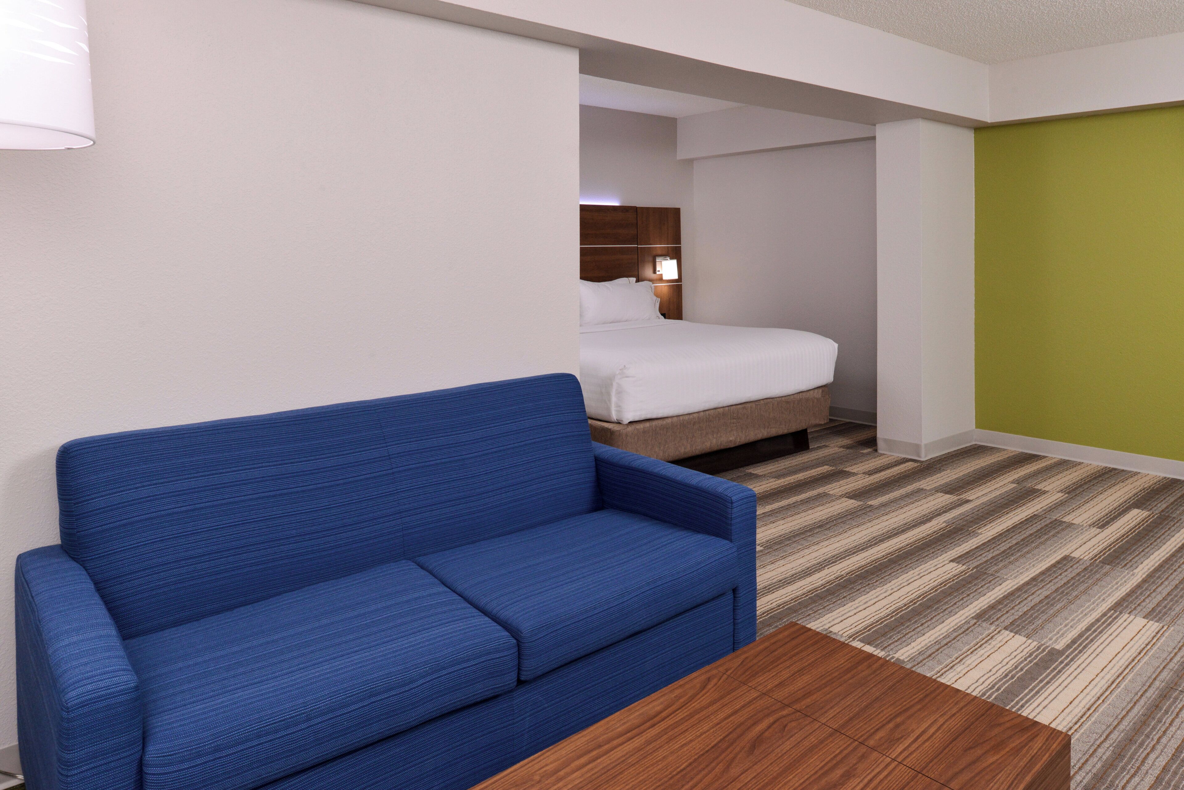 A hotel room with a blue sofa, a wooden coffee table, a bed with white linens, and a green accent wall.