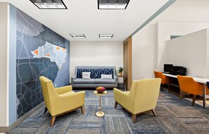 A modern hotel room with a sofa, two yellow armchairs, a small round table with a fruit bowl, a desk with a computer, and a map mural on the wall. - Holiday Inn Express & Suites Raleigh NE - Medical Ctr Area by IHG (Raleigh)