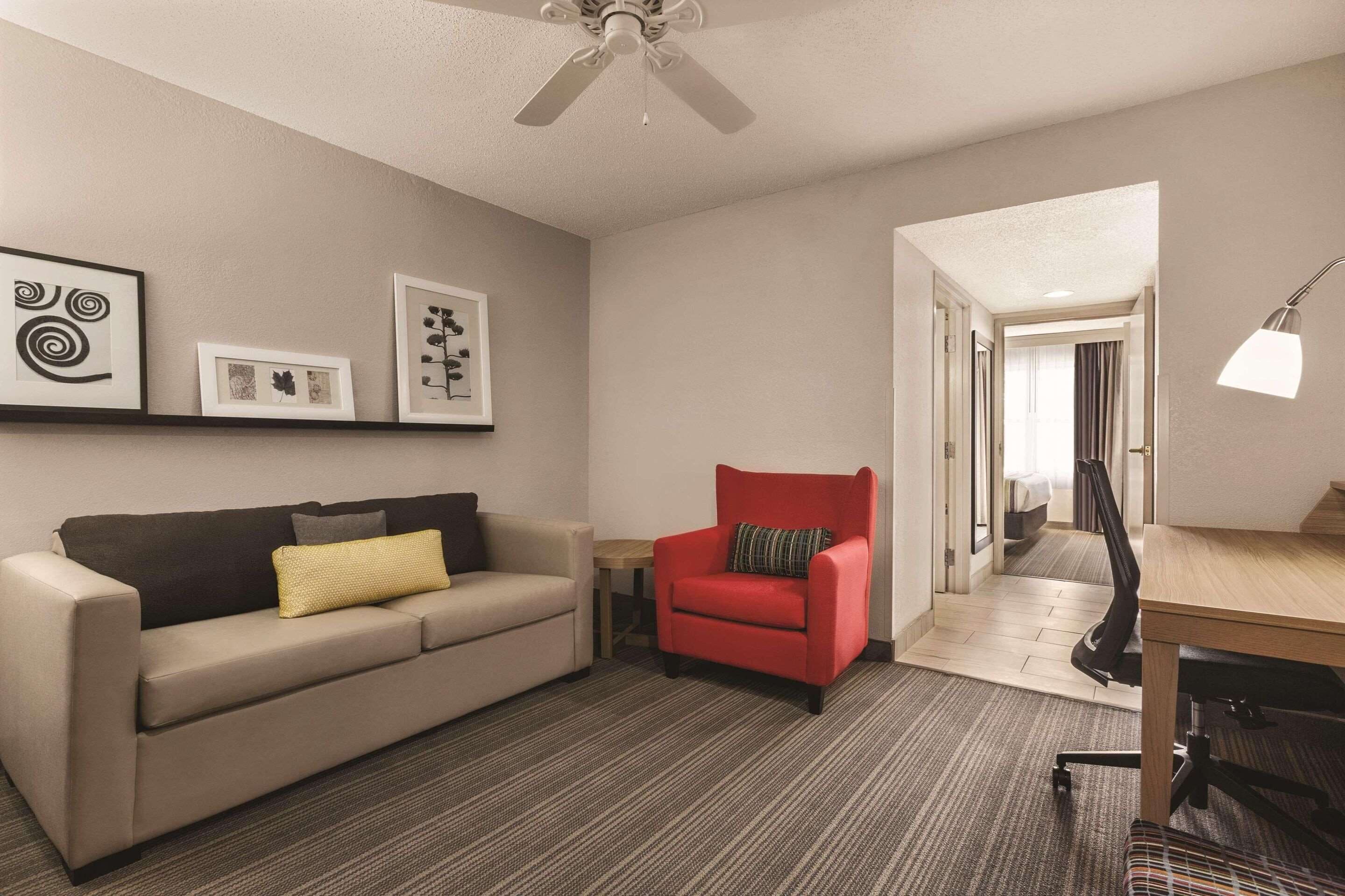 A hotel room with a sofa, a red armchair, a desk, and a ceiling fan.