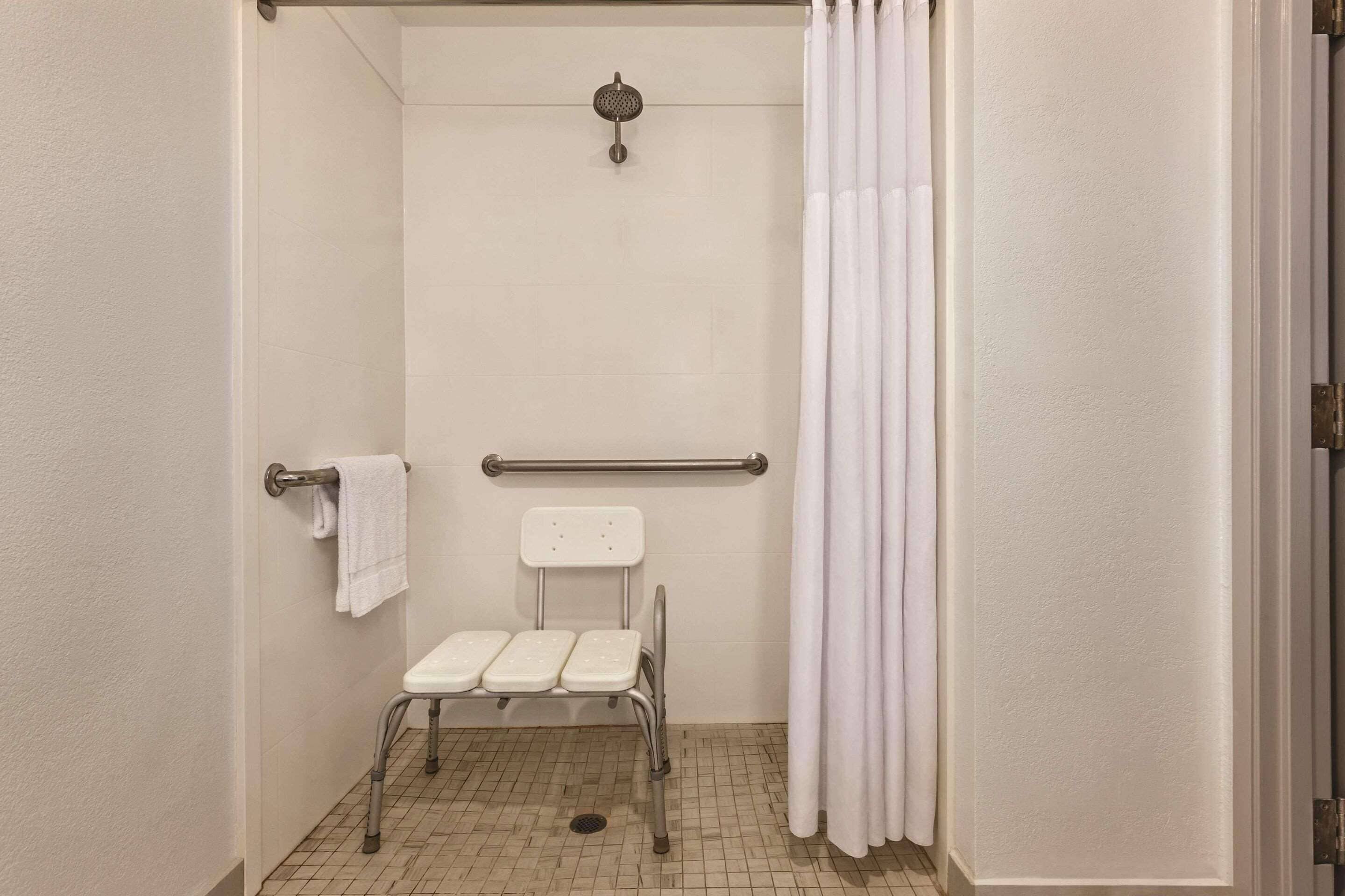 A bathroom with a white tiled floor, a white shower curtain, a grab bar, a white towel on a rack, and a white seat with armrests.
