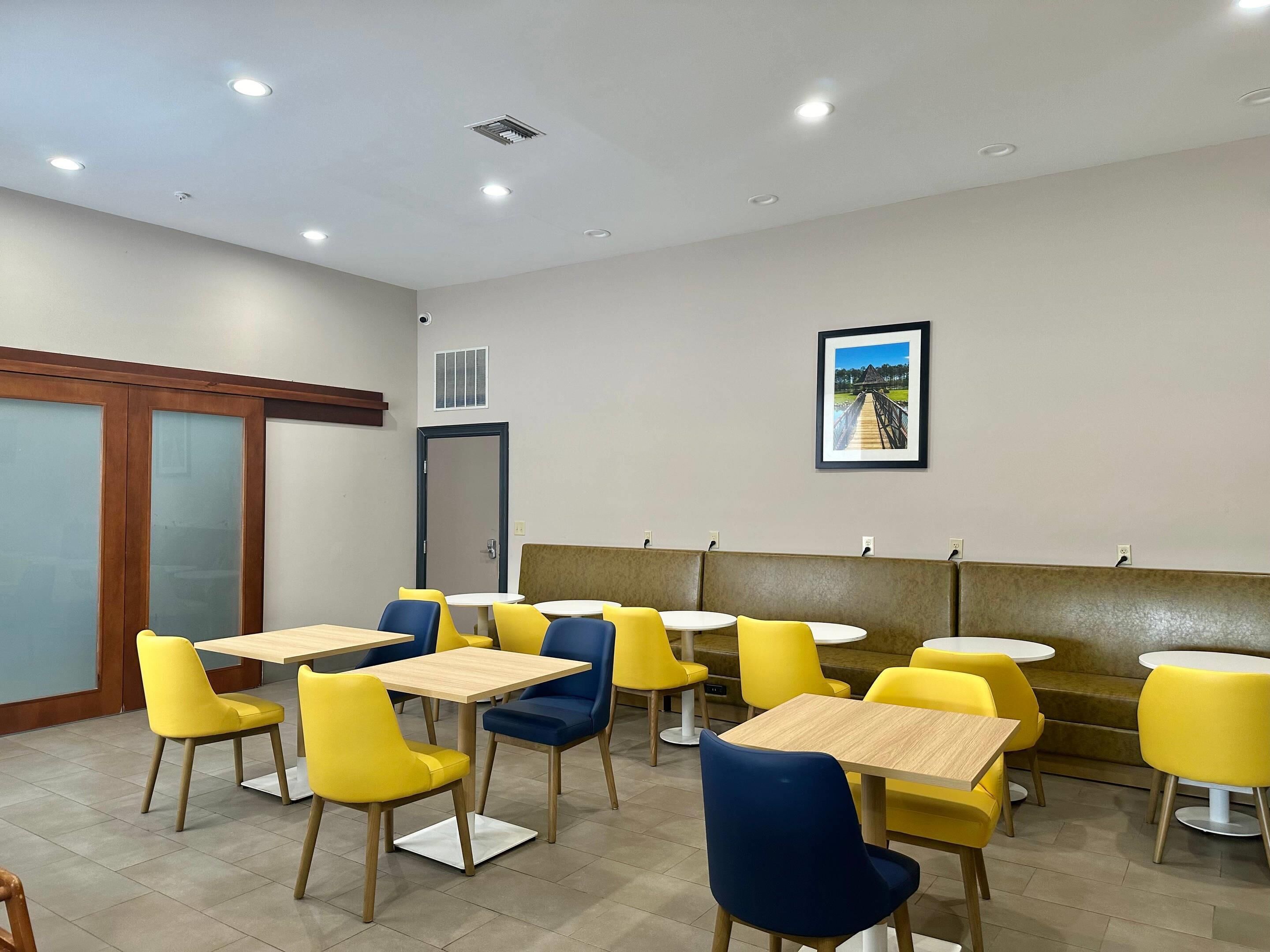 A dining area with yellow and blue chairs, wooden tables, a framed picture on the wall, and a door with a window.