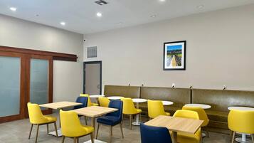 A dining area with yellow and blue chairs, wooden tables, a framed picture on the wall, and a door with a window.