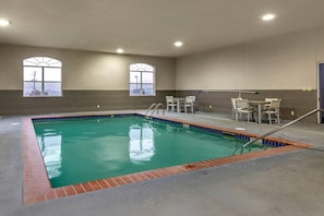 An indoor swimming pool with clear water, two windows with arched tops, and a seating area with chairs and a table.