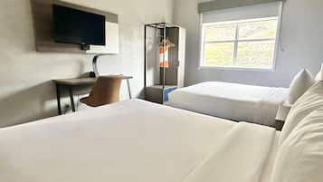 A hotel room with two beds, a desk, a chair, a TV, and a window with a view of greenery.