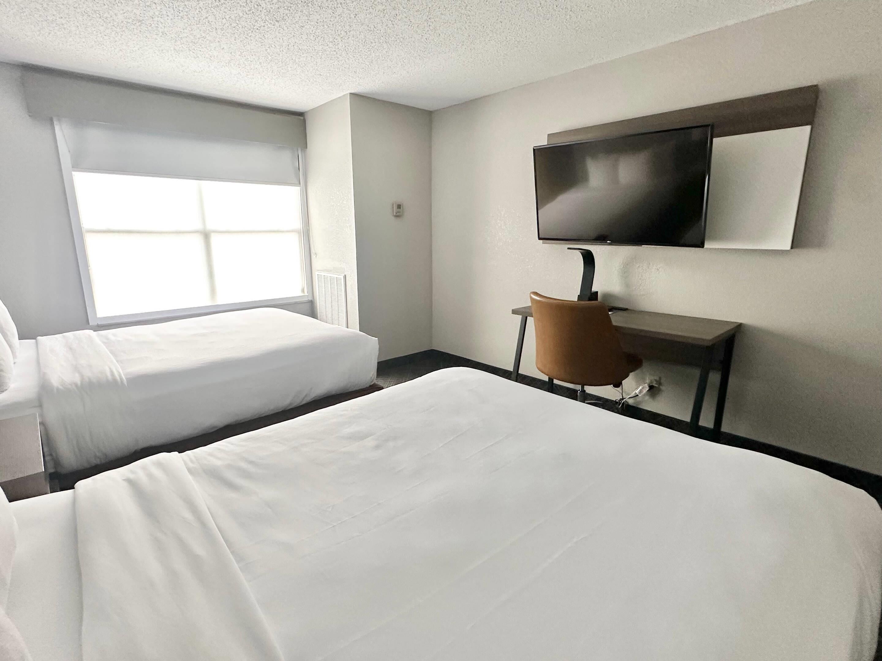 Quarto de hotel com duas camas, uma escrivaninha com cadeira, televisão e janela com persianas.