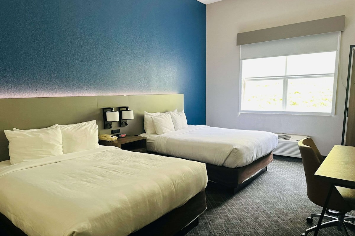 Une chambre d’hôtel avec deux lits, un bureau et une chaise. Il y a une fenêtre donnant sur un paysage verdoyant.